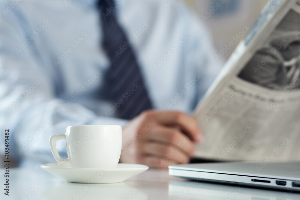 Cup of morning coffee on worktable with businessman reading news Cup of morning coffee on worktable with businessman reading news
