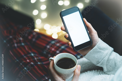 Girl holding smartphone with a empty blank screen monitor and a cup of coffee or tea on the background in a home atmosphere, hipster using in hands a smart phone with space for information, blur