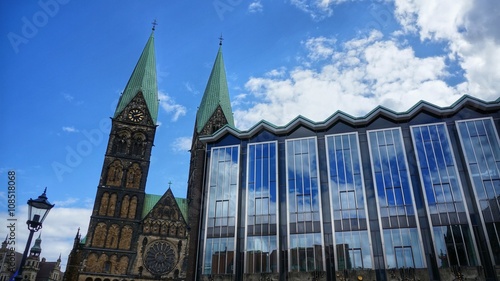 Bremen, Germany. Towers of Bremen Cathedral (German: 'Bremer Dom') and part of the Parliament of Bremen on a sunny day. Partly cloudy sky mirrored in the huge glass front of the government building.