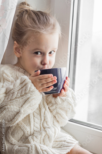 A little girl in the white knitted sweater sits on a window and drinks from a cup