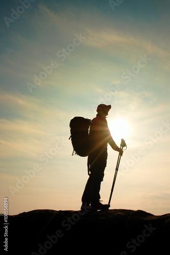 Hiker in windcheater, baseball cap and with trekking poles stand on mountain peak rock.