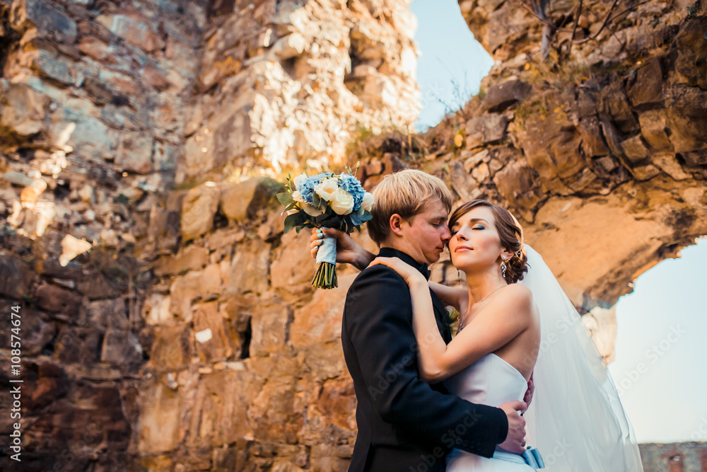 Handsome romantic groom kissing beautiful brunette bride near old wall castle