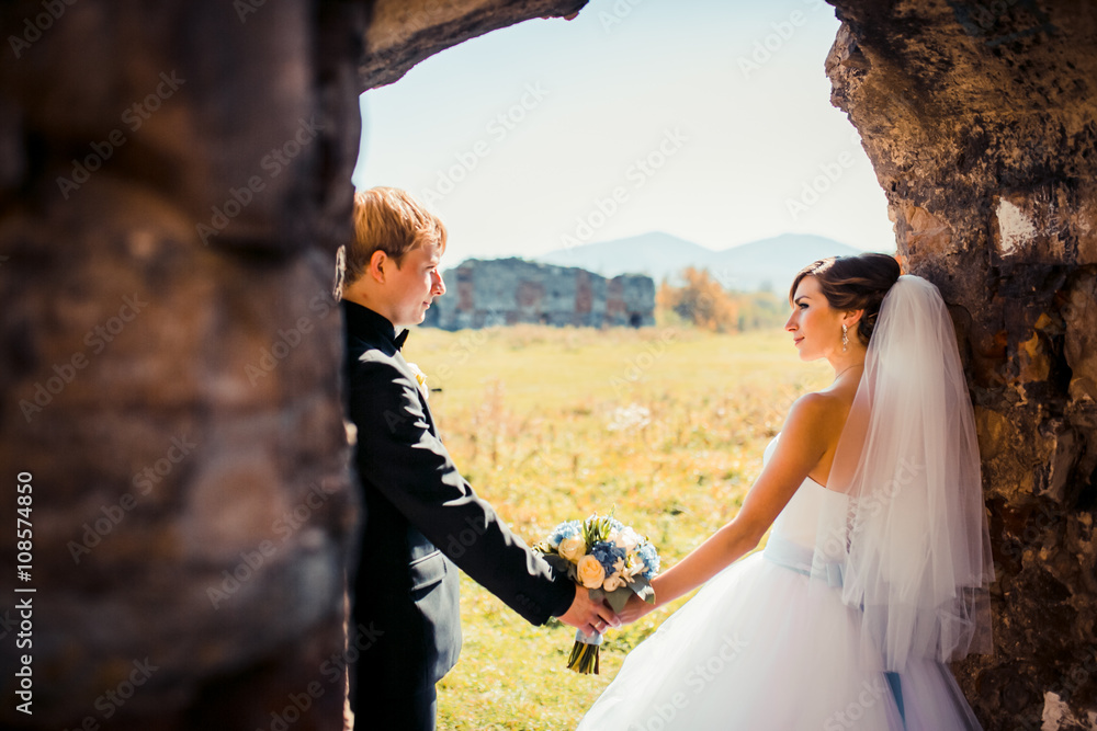 Handsome romantic groom kissing beautiful brunette bride near old wall castle