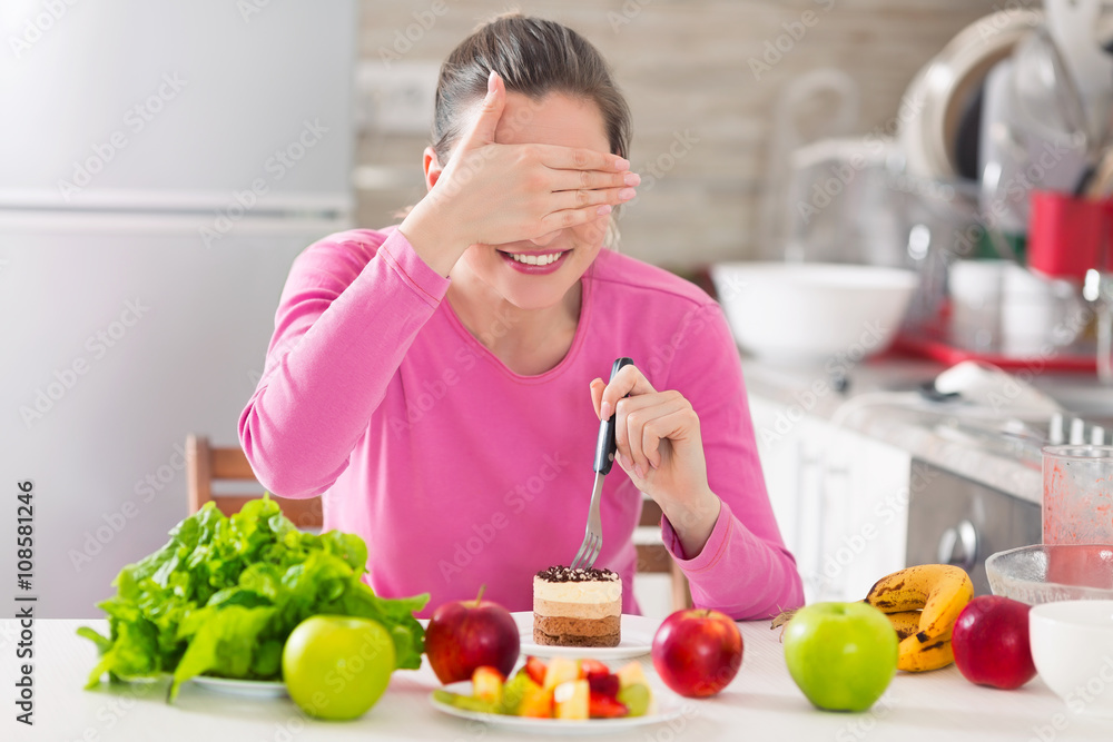 Young woman covering her eyes while eating a cake instead of healthy fresh fruit and vegetable on the table Young woman covering her eyes while eating a cake instead of healthy fresh fruit and vegetable on the table