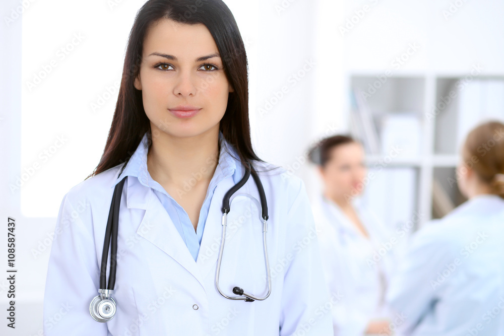 Brunette female doctor on the background of colleagues talking to each other in hospital Brunette female doctor on the background of colleagues talking to each other in hospital