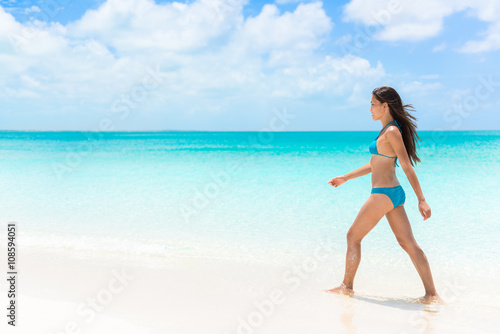 Beautiful bikini woman walking relaxing on sunny perfect white sand beach and pristine turquoise clear water in idyllic tropical destination during summer travel. Luxury getaway vacation.