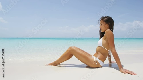 Beach body woman sun bathing on travel vacation. Sexy bikini body woman relaxing sun tanning lying down sunbathing on perfect paradise beach at tropical luxury destination in the Caribbean. 