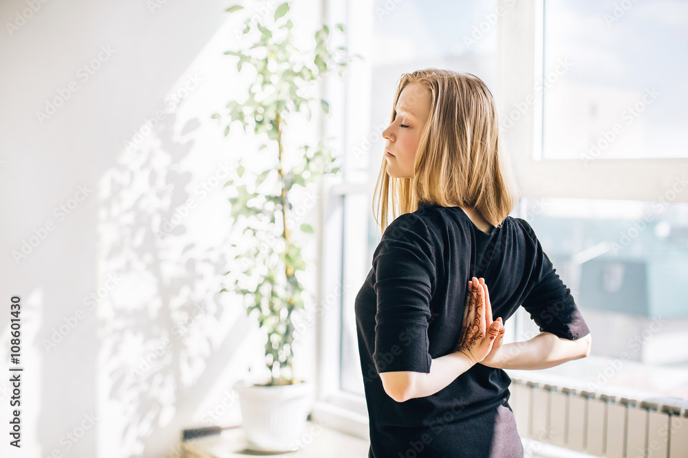 young blonde girl near the window doing meditation, hands in namaste behind the back