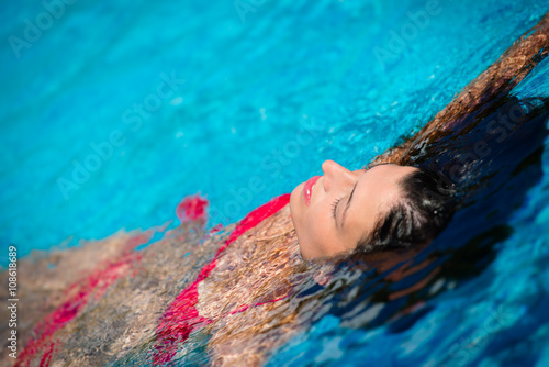 Young woman relaxing in resort swimming pool on summer vacation. Brunette caucasian model in red bikini floating in water.