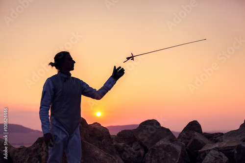 Fencer man wearing white fencing costume and throwing up his fencing sword on a background of sunset sky and rocks