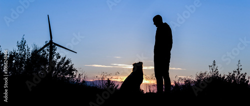 Silhouette von Hund und Herrchen von einem stimmungsvollen Abendhimmel in einer Hügeligen Landschaft mit Gräsern photo