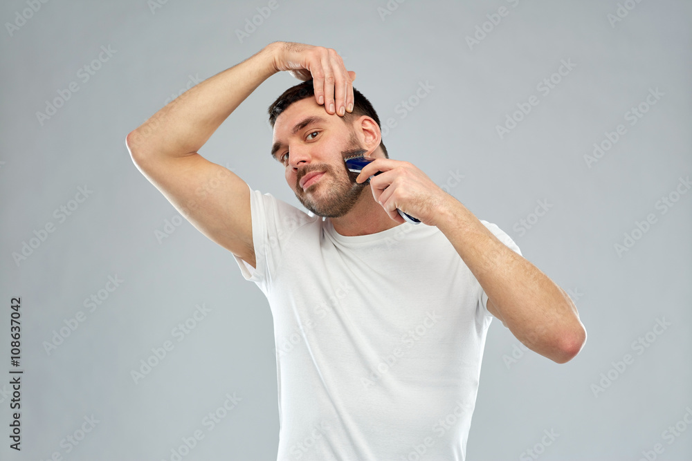Naklejka premium smiling man shaving beard with trimmer over gray