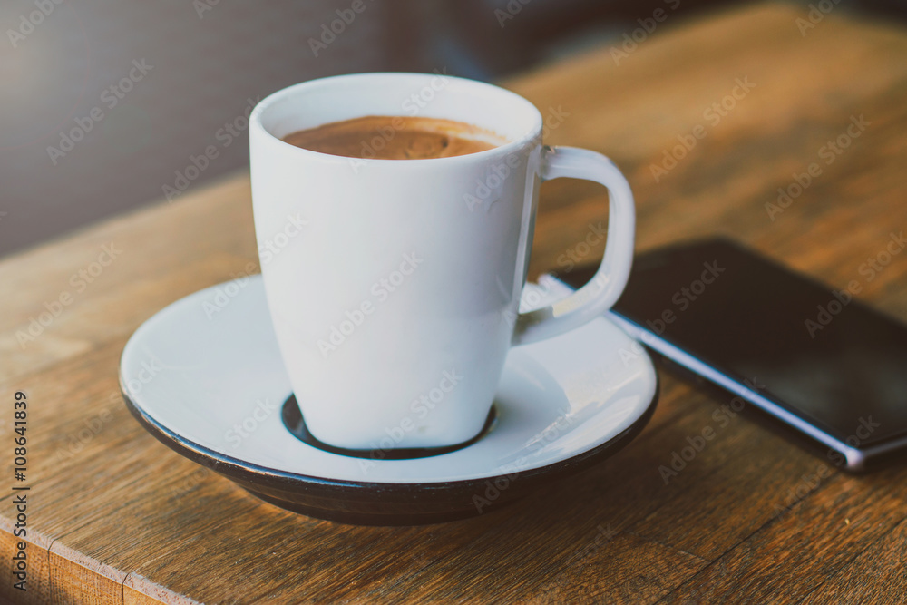 A cup of coffee and smart phone on a woden table in cafe, soft warm filter, lens flare