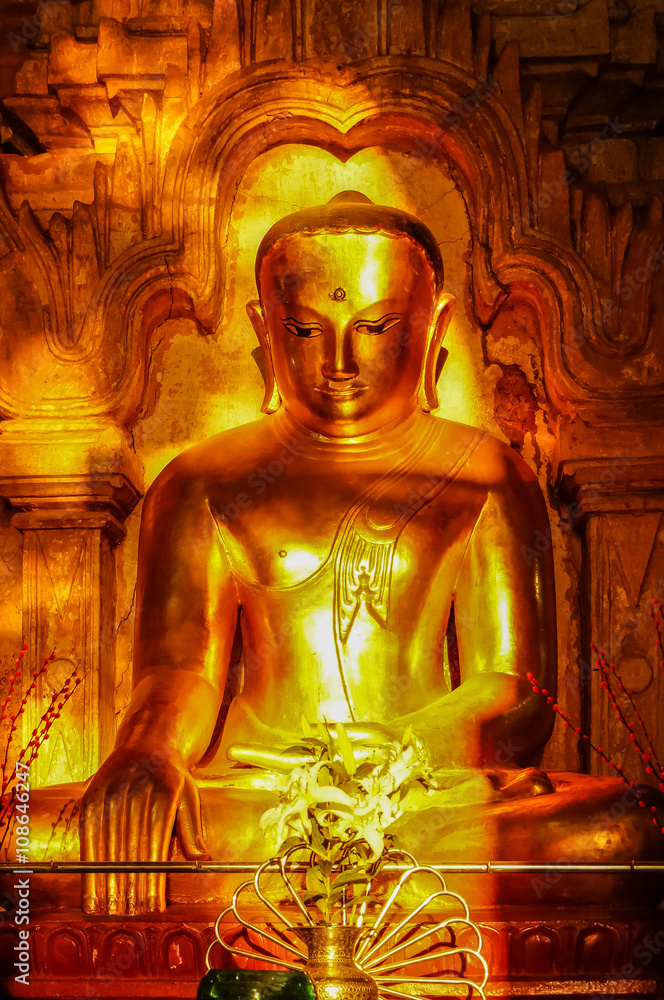 Antique golden sitting Buddha lit by late afternoon sun front view. Heavy gold colored Buddha with strong head and shoulders. The Buddha is seated in lotus posture typical for Theravada tradition.