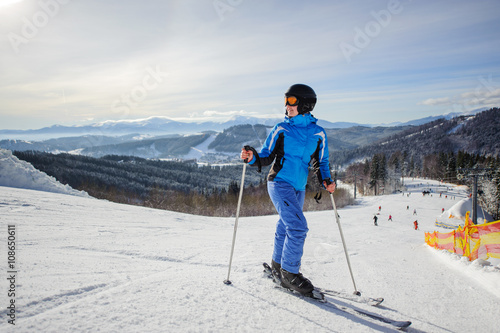 Young beautiful female skier on the middle of ski slope looking up. Girl at ski resort wearing helmet blue ski suit and goggles. Winter sports concept. Carpathian Mountains, Bukovel, Ukraine
