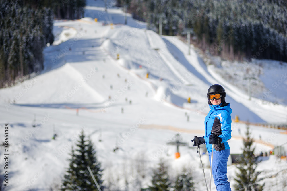 Young happy woman skier against ski slopes and ski-lift on background. Woman is wearing helmet skiing glasses gloves and blue ski suit. Winter sports concept.