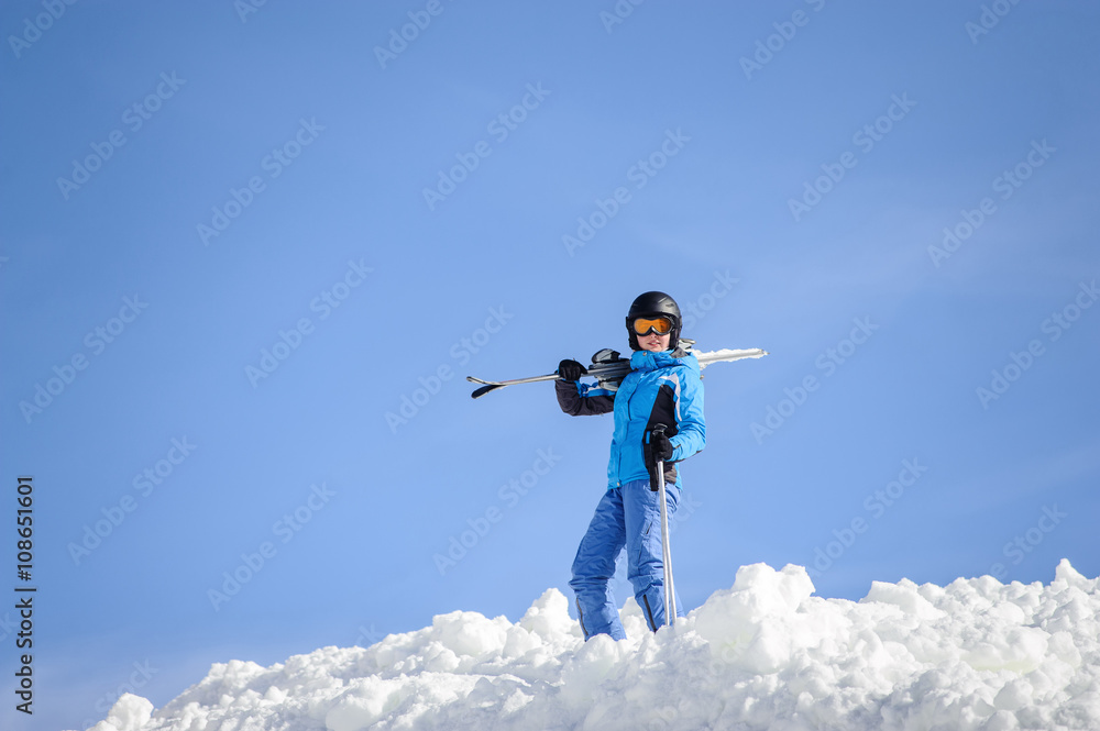 Portrait of young female skier standing on top of the mountain against blue sky on a sunny day. Woman is holding skis on her shoulder smiling and looking into the camera. Winter sports concept. Portrait of young female skier standing on top of the mountain against blue sky on a sunny day. Woman is holding skis on her shoulder smiling and looking into the camera. Winter sports concept.