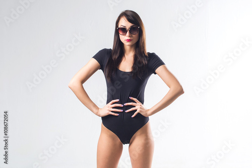 Glamorous female standing in studio, posing for portrait, wearing large sunglasses and one piece black bodysuit, holding her hands on waist, not isolated. © undrey