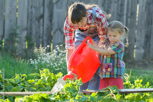 Woman gardener helping her daughter to pour vegetable garden bed with cucumbers