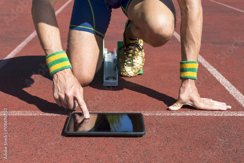 Athlete in gold running shoes crouching at the starting line of a running track wearing Brazil colors wristbands using his tablet