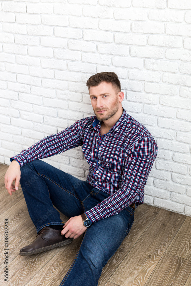 Handsome young man smiling while sitting on the floor and leaning at the wall