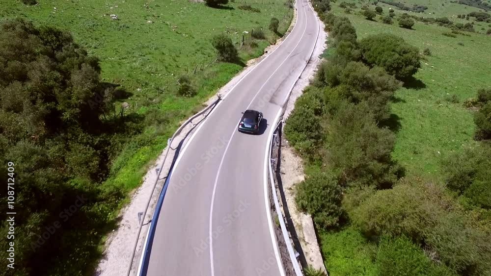 Camera follows with distance from above the lonely driving car on the road. Right and left of the street is green and hilly terrain without a building. Camera follows with distance from above the lonely driving car on the road. Right and left of the street is green and hilly terrain without a building.
