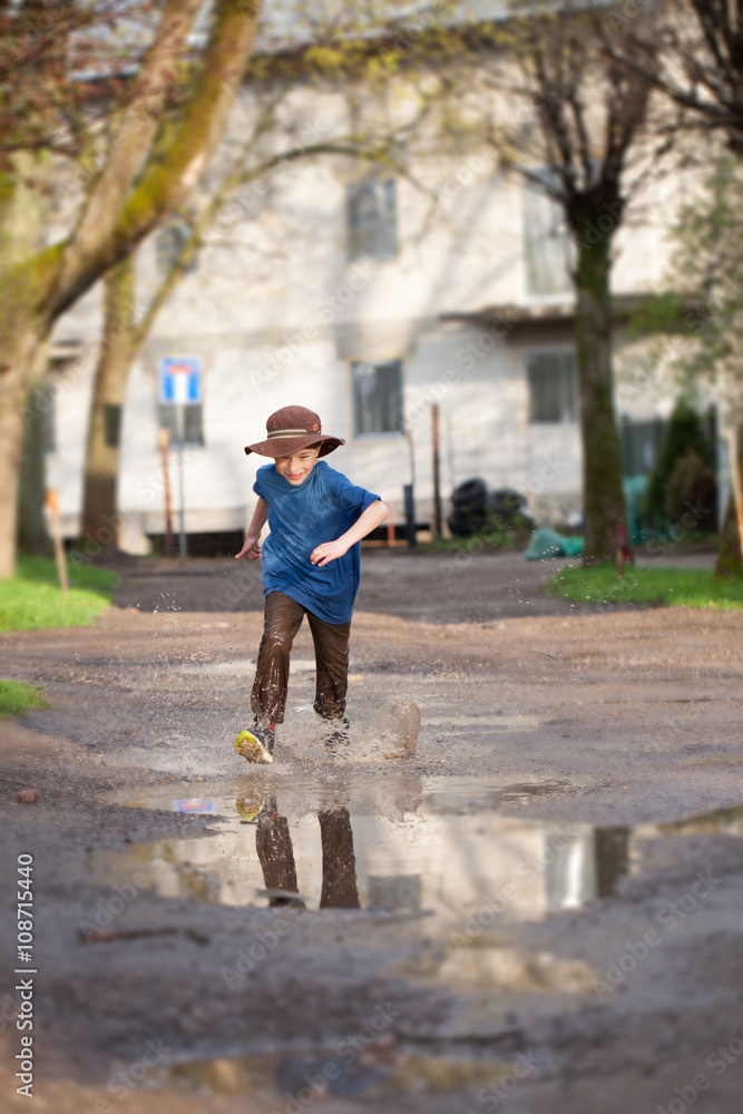 Fototapeta premium Little boy splashing in a mud puddle, jumping into a puddle 