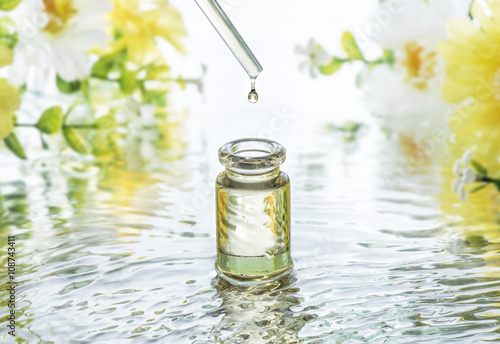 The bottle of Moisturizing cosmetic oil in the water waves on the summer flowers blur background and  pipette with oil drop above the bottle photo