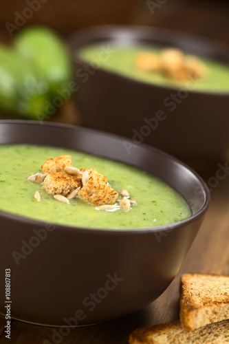 Zucchini cream soup with wholewheat croutons and roasted sunflower seeds served in brown bowl with toastbread on the side (Selective Focus, Focus on the front of the croutons)