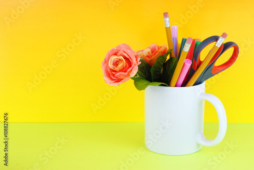 White coffee mug filled with markers, pencils, scissors and a silk rose on a green and yellow background good for secretary's day, or administrative professionals day