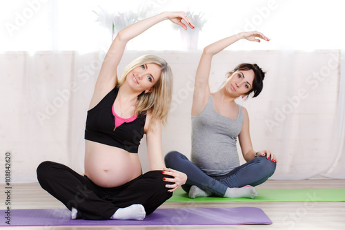 Pregnant cute women,blonde and brunette,dressed in sports attire, perform stretching exercises in prenatal yoga to strengthen the muscles that hold classes in the gym bright in the daytime