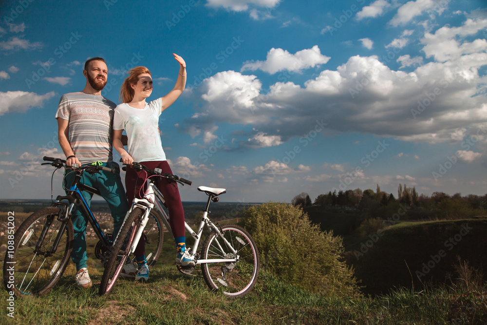 Obraz premium Beautiful young couple in love walking with bicycles, looking towards the beautiful nature and the blue sky. The concept of active rest