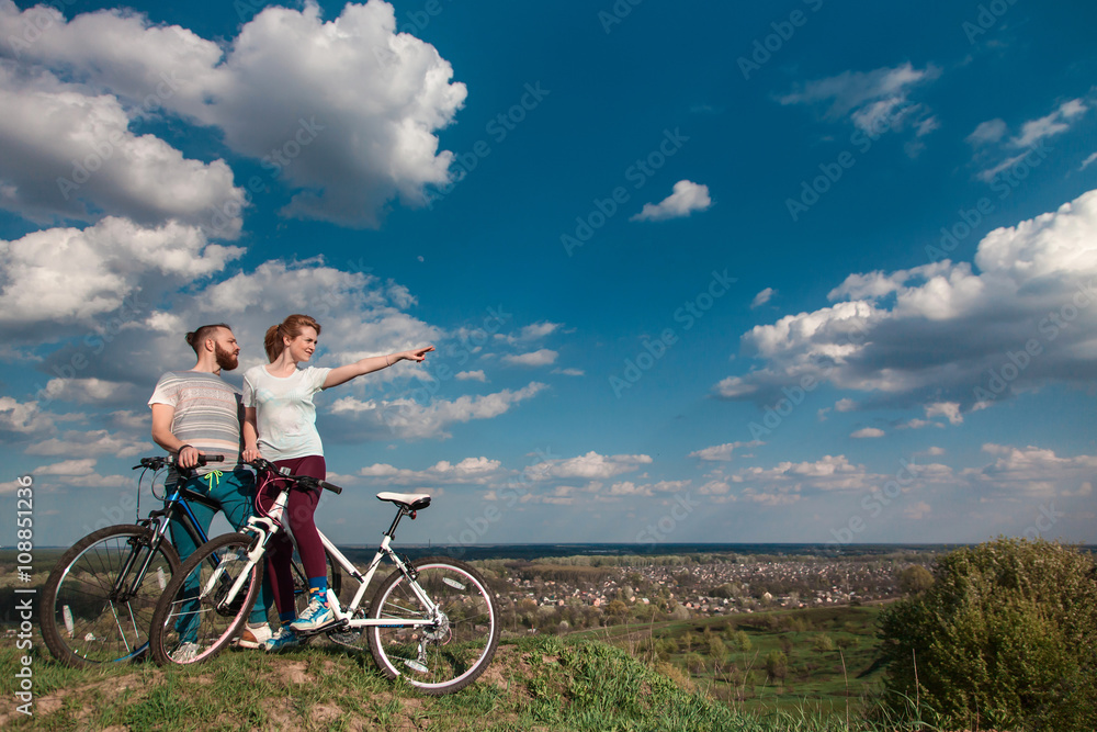 Obraz premium Beautiful young couple in love walking with bicycles, looking towards the beautiful nature and the blue sky. The concept of active rest