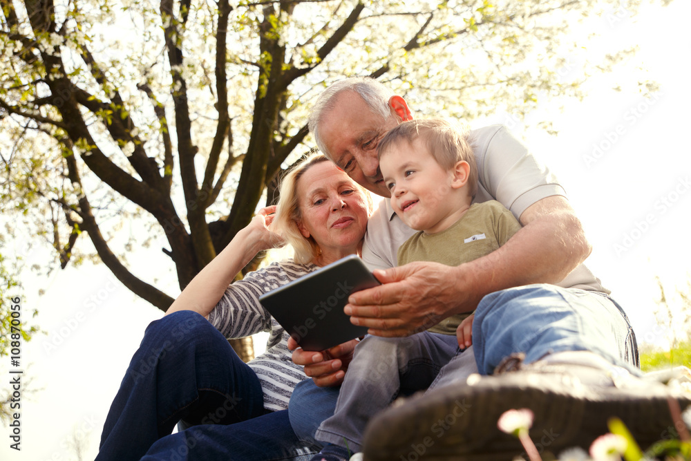 Grandparents with grandson enjoying the sunny spring day outdoors. They are looking something on tablet.