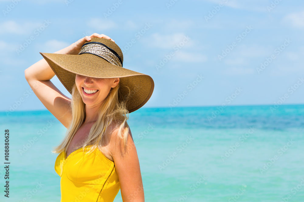 Carefree beautiful fashion blonde woman in beach straw hat and long yellow dress flying in the wind at the tropical beach seashore. Natural woman beauty. Lady hold her hat and smile to the camera.