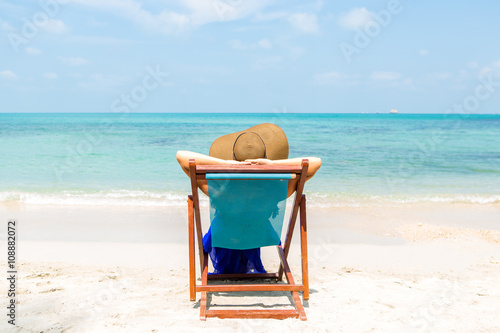 Beautiful mammy in long blue dress and beach straw hat relaxing in a beach chair. Cute woman relaxing and look to the sea shore. Lady near coastline. Photo from back. Happy mothers day.