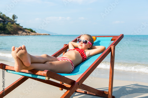 Cute little child in red swimsuit and white sunglasses enjoying on a sun lounger rest on the beach chair on tropical sandy beach sea shore. Sunbathing and leisure on sunny day.