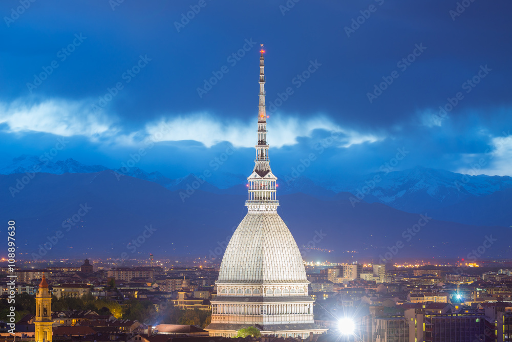 Cityscape of Torino (Turin, Italy) at dusk with details of the Mole Antonelliana towering on the city and glowing in the night. Wind storm over the Alps in the background.