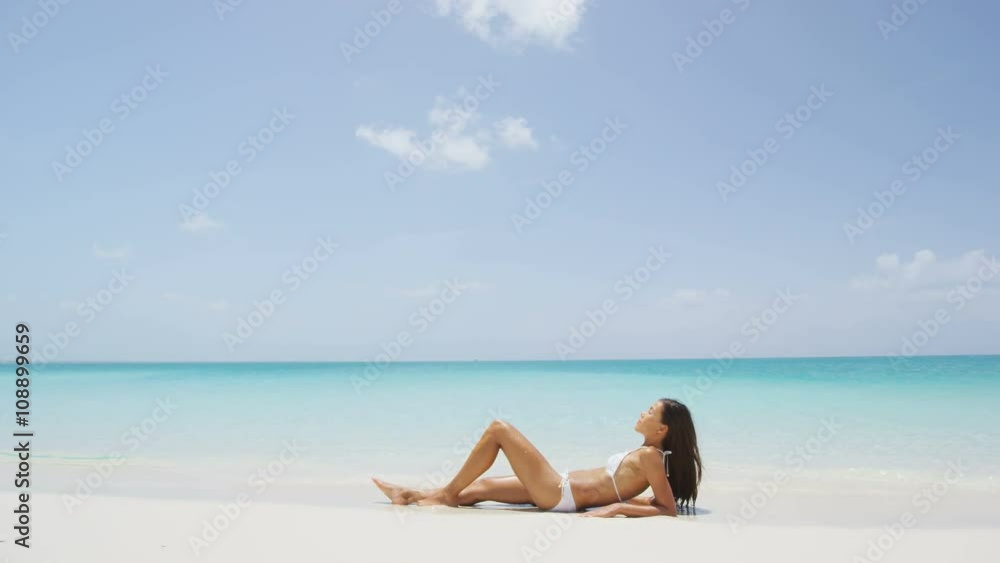 Suntan woman relaxing on summer beach vacation sunbathing. Sexy young ethnic adult in white bikini lying down on sand with sky copyspace for tropical travel destination concepts. SLOW MOTION. Suntan woman relaxing on summer beach vacation sunbathing. Sexy young ethnic adult in white bikini lying down on sand with sky copyspace for tropical travel destination concepts. SLOW MOTION.