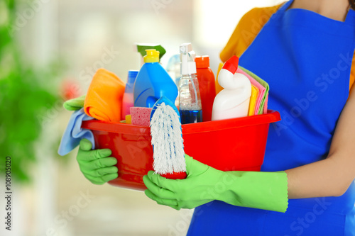 Woman in rubber gloves holding basin with detergents on blurred background