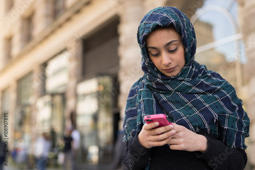 Young woman wearing hijab in city texting on cell phone photo