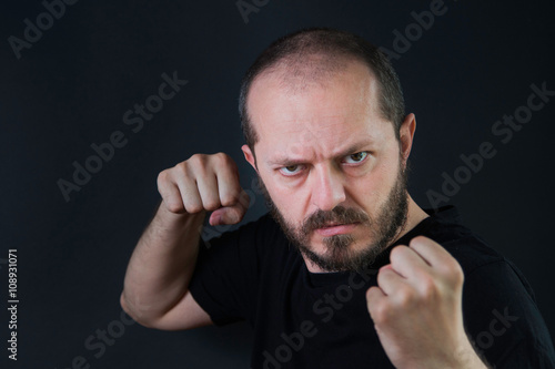  Serious aggressive man with beard and mustaches on black background in low key, holding fists and threatening, ready for fight 