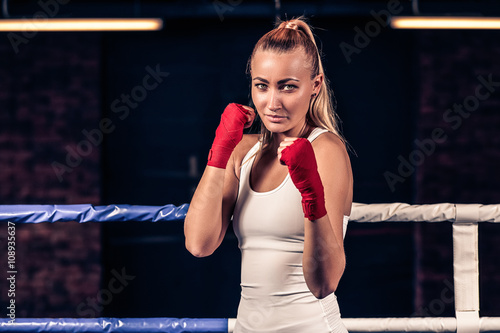 Portrait of young beautiful boxer woman ready to kick with red boxing bandage on hands in gym. Strong hand and fist, ready for fight and active exercise.