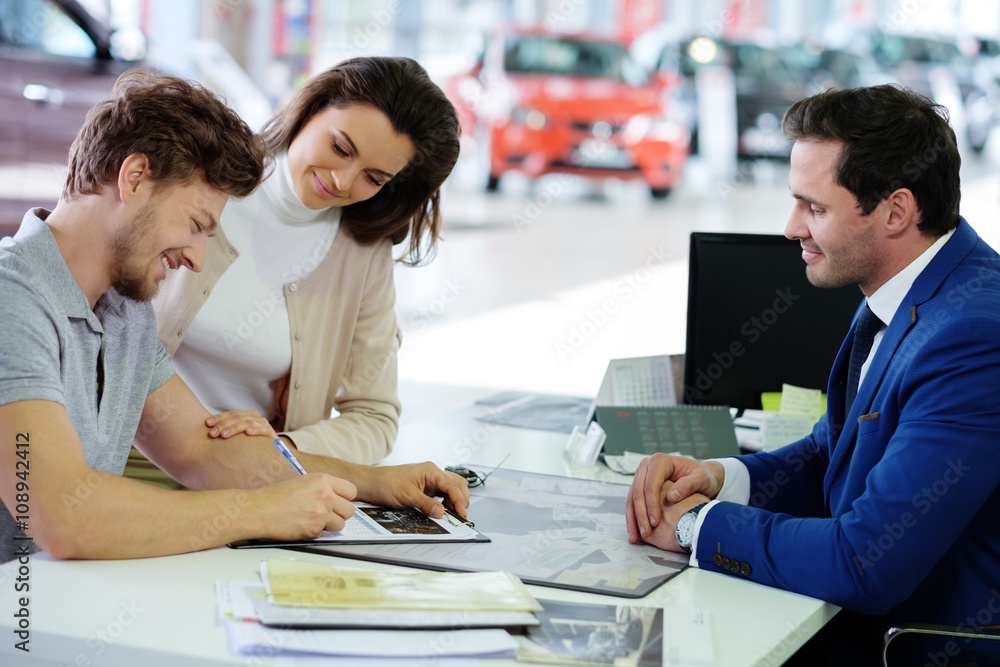 Fototapeta premium Happy young couple signing a contract to purchase a new car at the dealership showroom.