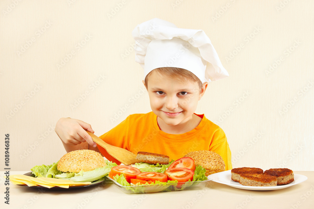 Young smiling boy in chef hat puts meat on the hamburger Young smiling boy in chef hat puts meat on the hamburger