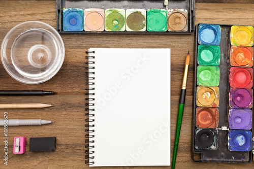 Workstation concept top view : Water color,brush,pen,marker,pencil,rubber,sharpener,glass of water and color pencil on wooden background