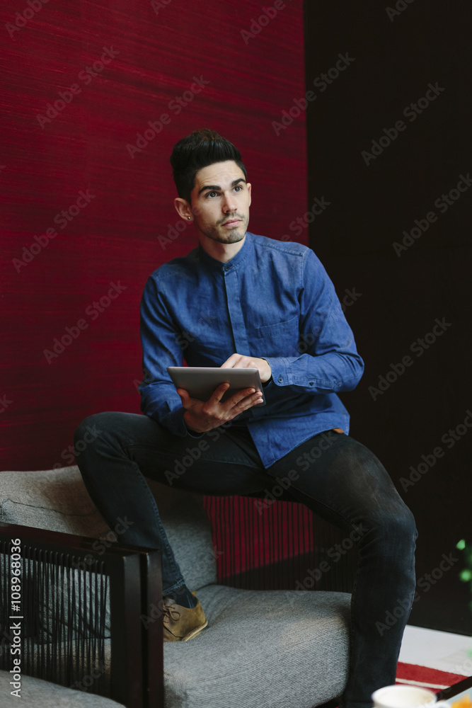 Young businessman sitting on armchair of a lobby with his digital tablet watching something Young businessman sitting on armchair of a lobby with his digital tablet watching something