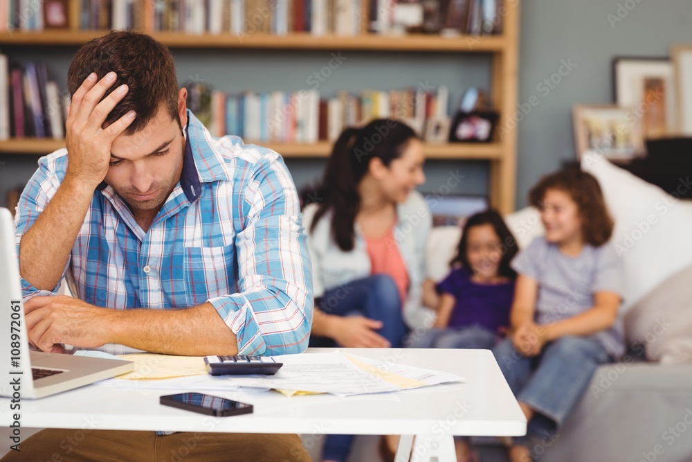 Tensed man by laptop while family sitting in background Tensed man by laptop while family sitting in background