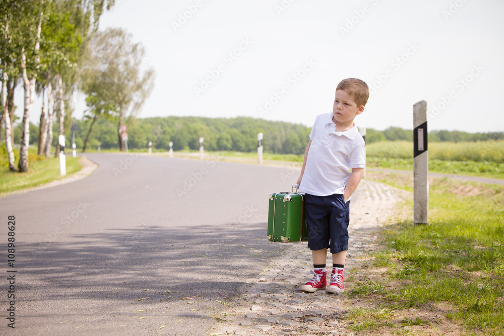 Adorable little kid boy in a white polo shirt and shorts holds a green retro suitcase and stands on the countryside road waiting for a bus or a car. Ready for travel. Vacation time. Child on the road Adorable little kid boy in a white polo shirt and shorts holds a green retro suitcase and stands on the countryside road waiting for a bus or a car. Ready for travel. Vacation time. Child on the road