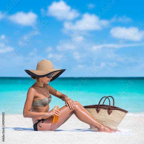 Sun protection uv rays skincare. Sunscreen spray bottle woman applying body lotion on smooth legs. Girl tanning putting sunblock Beach essentials for summer holidays: straw hat, sunglasses, tote bag.